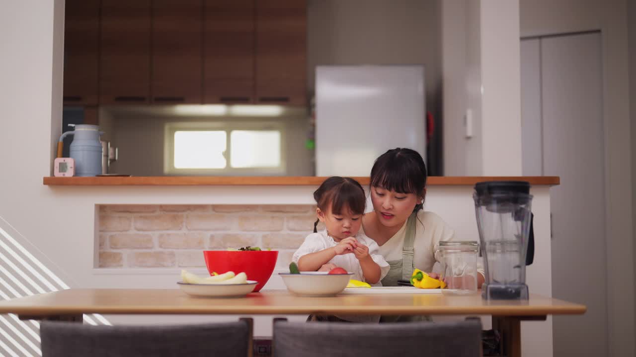 母亲和她的小女儿在家里准备沙拉。妈妈帮她的小女儿准备蔬菜，鼓励她健康饮食视频素材