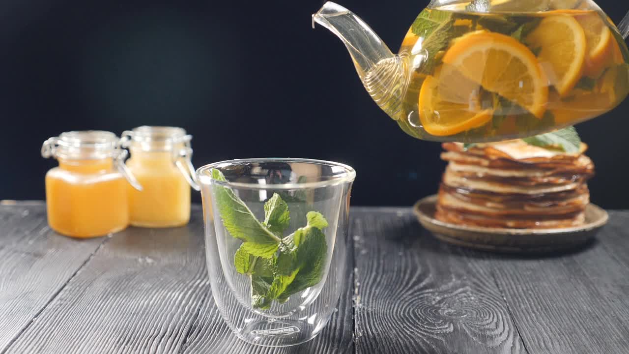 将橙汁、柠檬汁、青柠汁从透明的茶壶中倒入装有九香脂的玻璃杯中。茶具、玻璃茶壶及茶杯。以蜂蜜和薄饼为背景泡茶。慢镜头茶。全高清视频下载