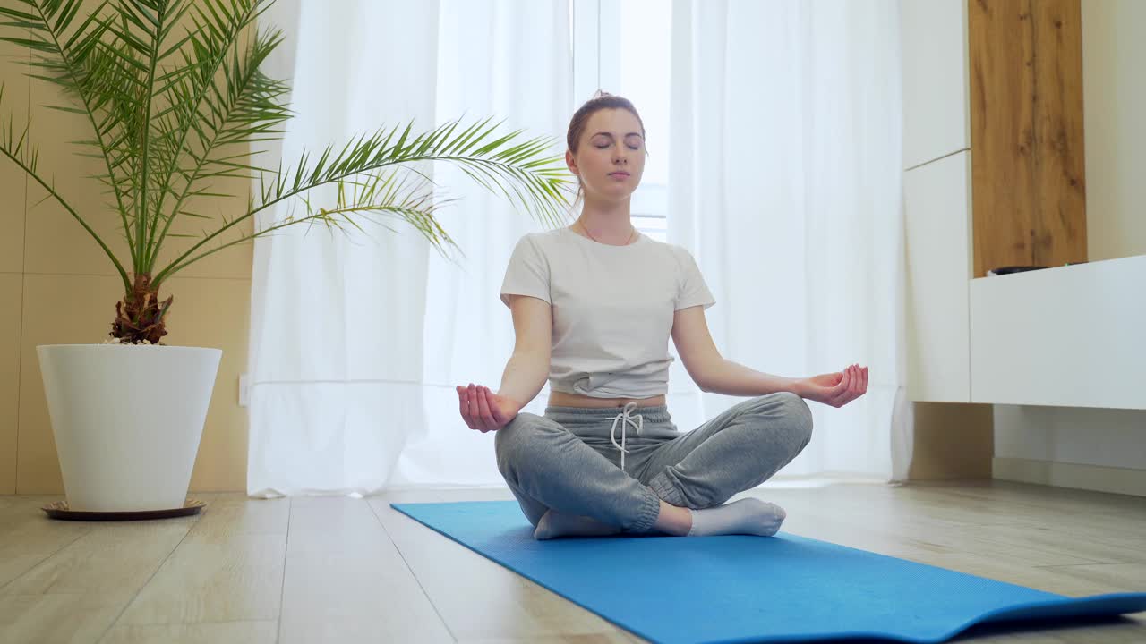 一个年轻的红发女孩在家里练习瑜伽。活跃的女人穿着便服坐在客厅的垫子上放松和冥想视频素材