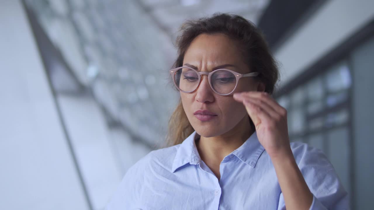 劳累过度的西班牙妇女摘下眼镜触摸疼痛的太阳穴，感到头痛，在大厅休息忍受偏头痛，天气依赖性疼痛，经前综合症，疲惫视频素材