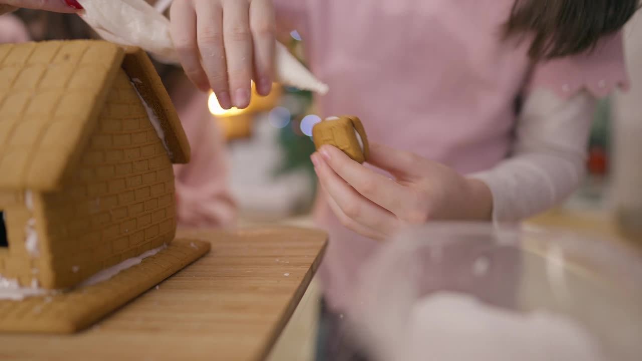 现场镜头跟随孩子们手中的姜饼，涂上糖胶，把糕点放在房子上。一个陌生的白人女孩在新年和妈妈一起做传统的甜点视频素材