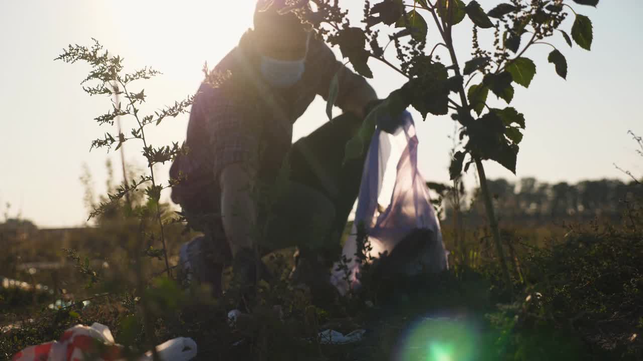 戴手套的男性生态活动家在农村用袋子收集垃圾。年轻的义工戴着口罩清理路边的废纸草坪，拯救大自然。环境问题的概念。慢动作视频素材