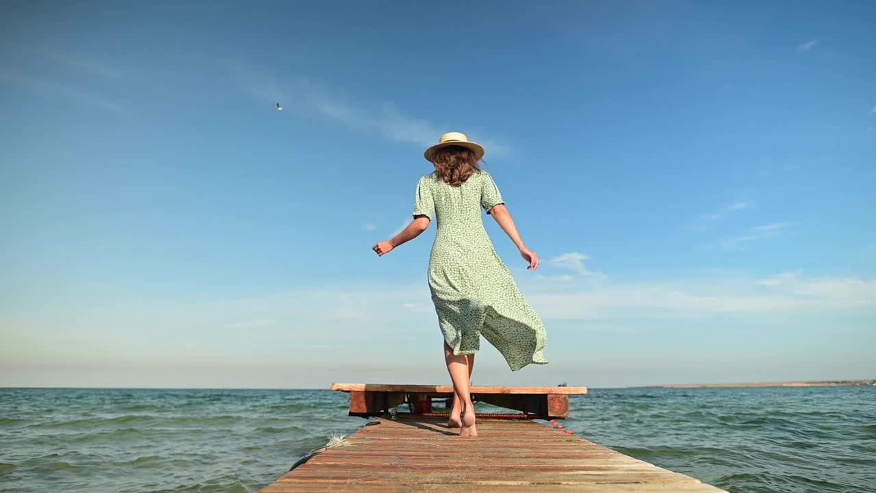 一个身穿绿色夏装、头戴草帽的年轻女子，在风的吹拂下，在清晨的海上沿着木码头缓缓走着。行走优雅，美丽和女性气质视频素材