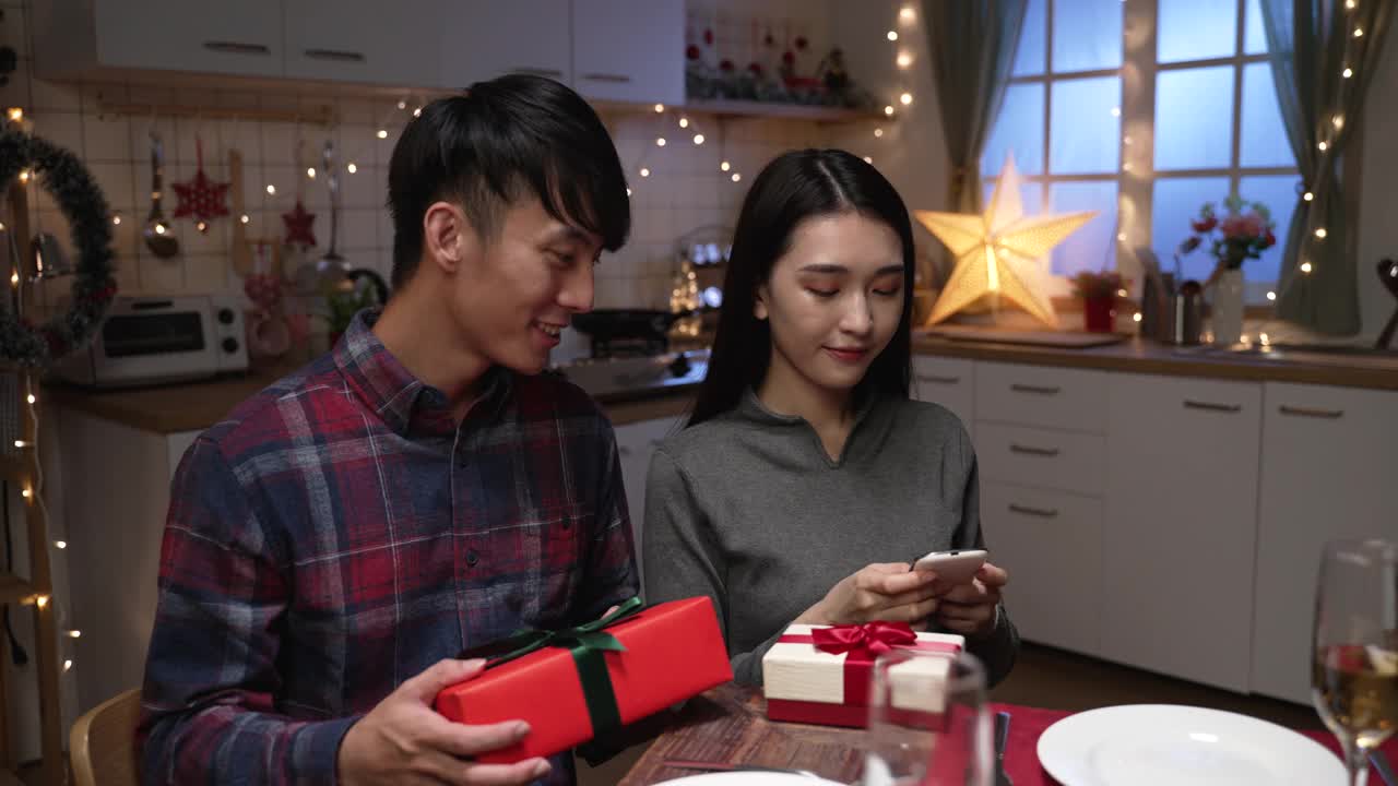 在情人节的约会之夜，兴奋的年轻夫妇坐在厨房的餐桌旁交换礼品盒。两个恋人拿着礼物和手机一起自拍视频素材