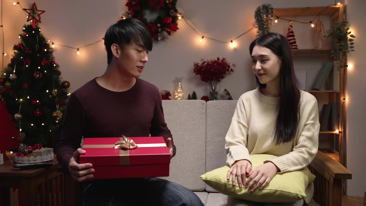 年轻幸福的夫妇放松在舒适的沙发在圣诞节假期。男人坐在圣诞树旁的沙发上给女朋友送礼物。愉快的女士满意的圣诞礼物在室内视频素材