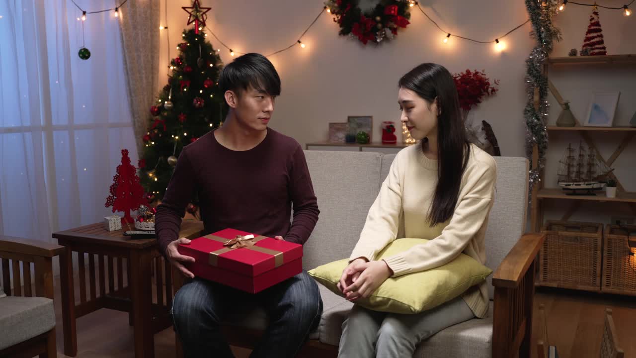 漂亮的年轻夫妇正在家里庆祝圣诞夜。漂亮女孩满意地收到来自她的男朋友坐在家里的沙发上的礼盒。圣诞节晚上，男人给妻子送礼物。视频素材
