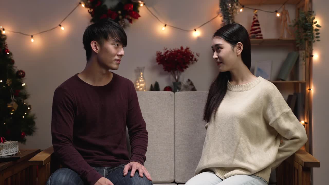 在圣诞前夜，相爱的年轻夫妇在家里一起庆祝圣诞节。亚洲男人惊讶于女友的礼物。女人坐在沙发上给震惊的男友送礼物。视频素材