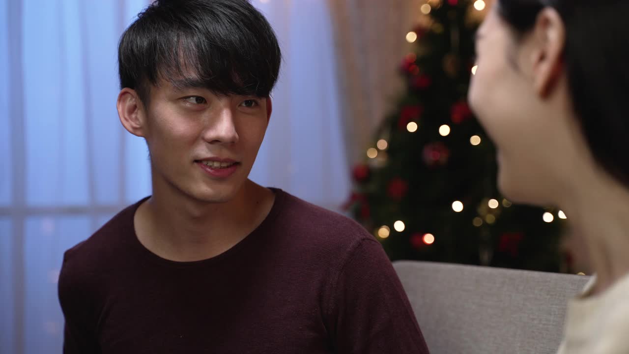漂亮的亚洲年轻夫妇坐在沙发上在家庆祝圣诞节。害羞的帅哥微笑着送给女友礼盒。惊喜的女孩收到礼物，愉快地笑着坐在沙发上视频素材