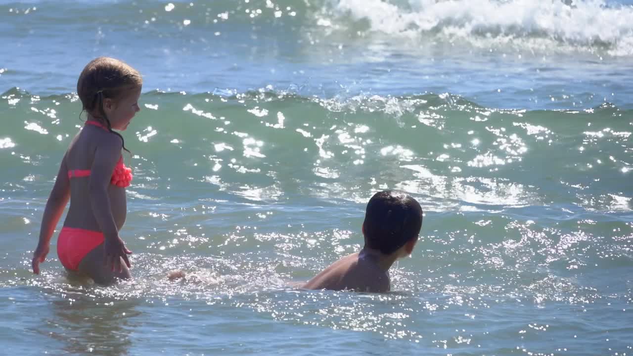 一个快乐的小男孩和一个可爱的小女孩穿着泳衣在海浪中跳跃视频素材