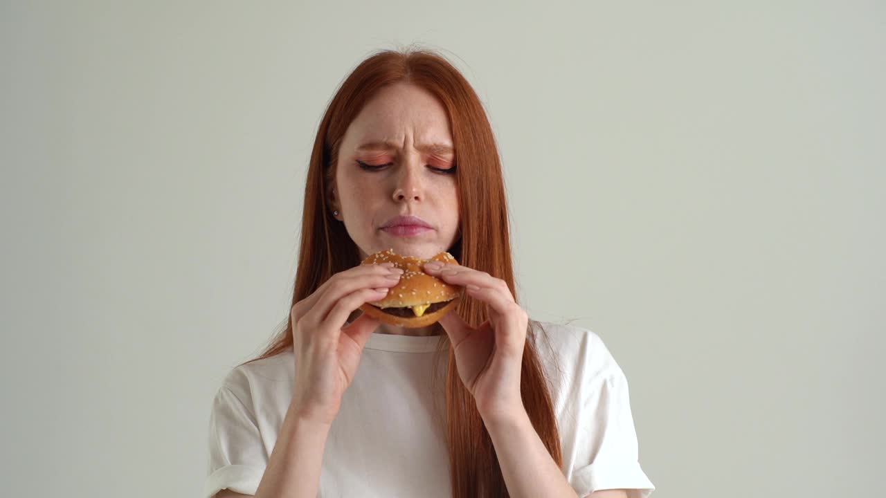 愉快的年轻女子的特写照片，胃口吃美味的汉堡和看着相机站在白色孤立的背景在工作室视频素材