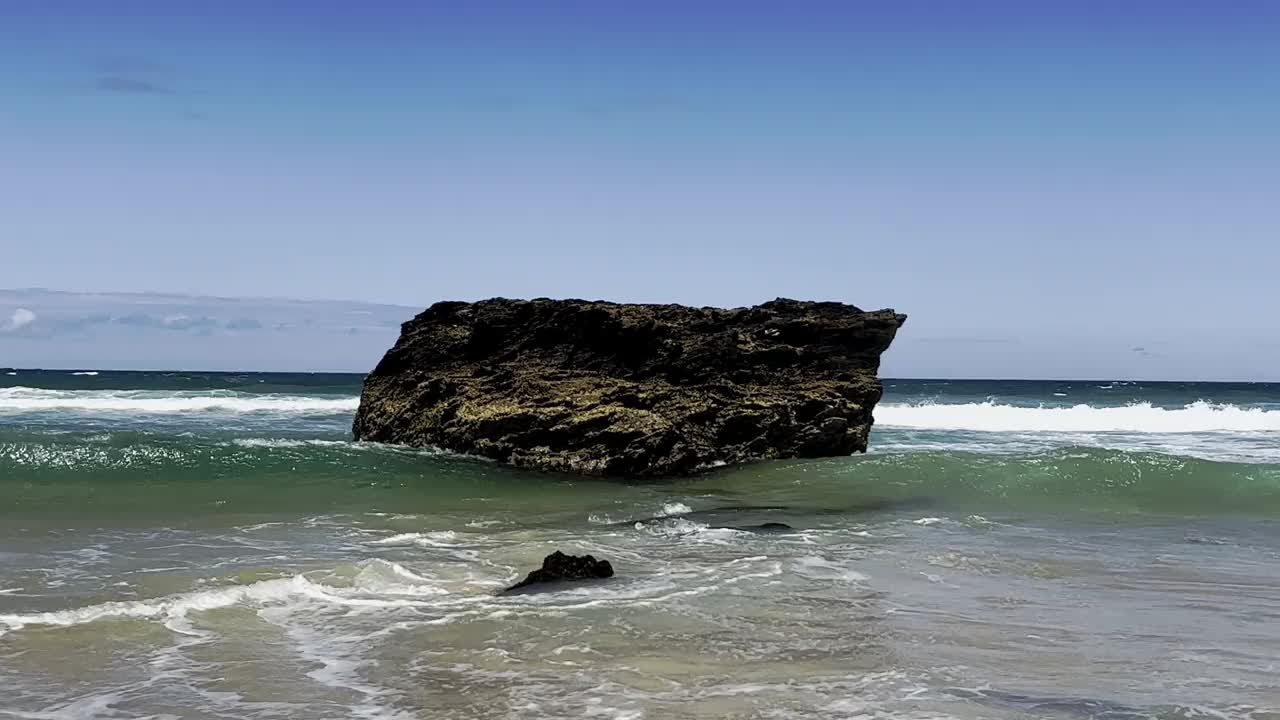 在沙滩上的冲浪区，一块巨大的巨石受到持续的海浪作用的影响。视频素材