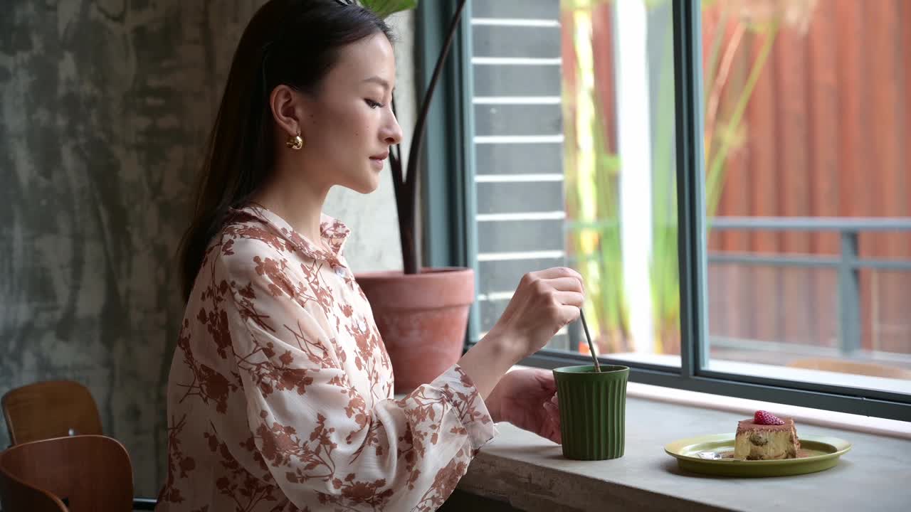 漂亮的中国亚洲年轻女子戴眼镜，喝一杯拿铁咖啡在咖啡馆的窗口，看着它。休息时放松。在咖啡店等待的年轻学生或女商人。视频下载