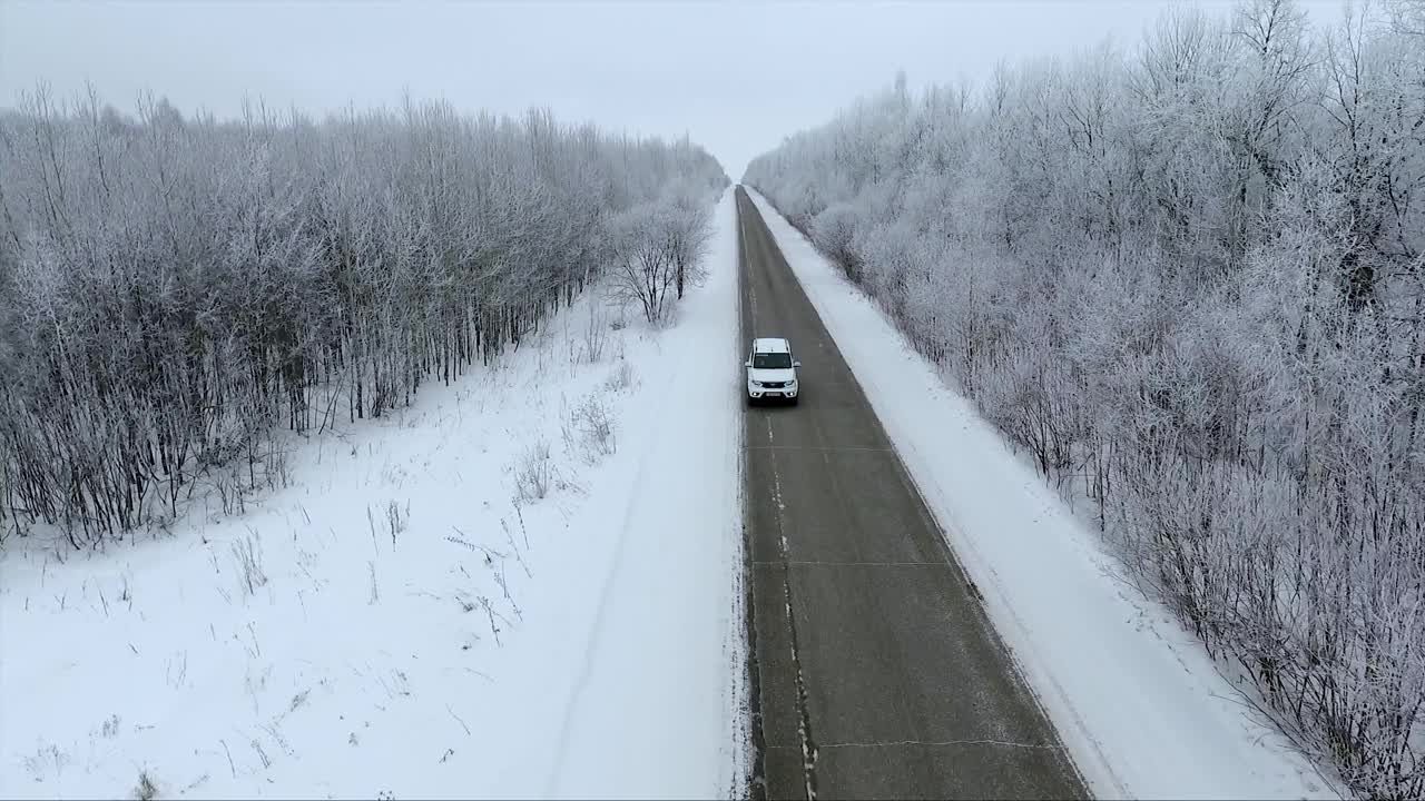 用无人机拍摄的4K视频显示，一辆白色汽车在冬天沿着积雪覆盖的树木之间笔直的道路行驶。视频素材