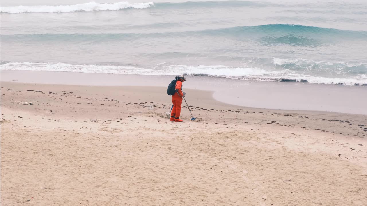 一个穿着红色衣服，带着金属探测器的男人沿着海滩走。慢镜头，4k，慢镜头视频素材