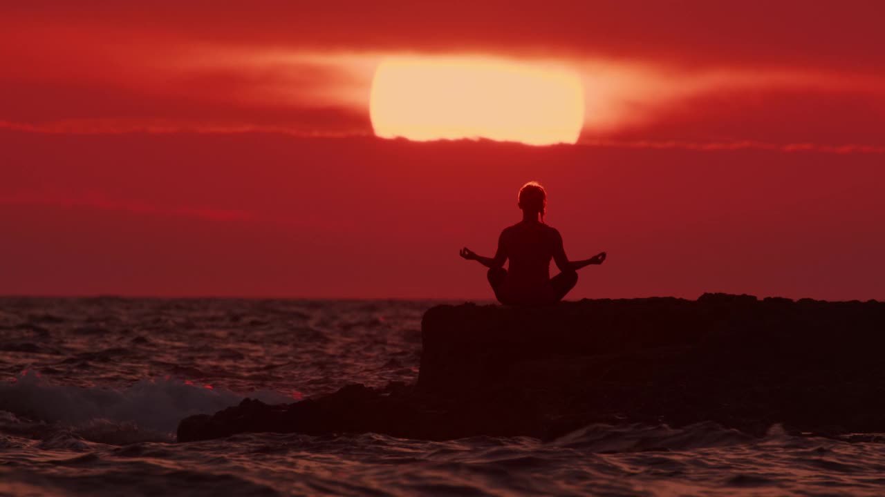 一个女人的剪影，在夕阳红天空的背景下沉思，坐在海洋或大海的岩石海岸上。人与自然和谐相处视频素材