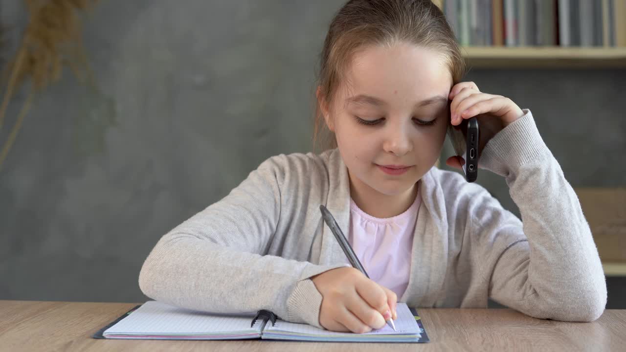 一个女学生坐在家里的桌子边打电话，在笔记本上做笔记。视频素材