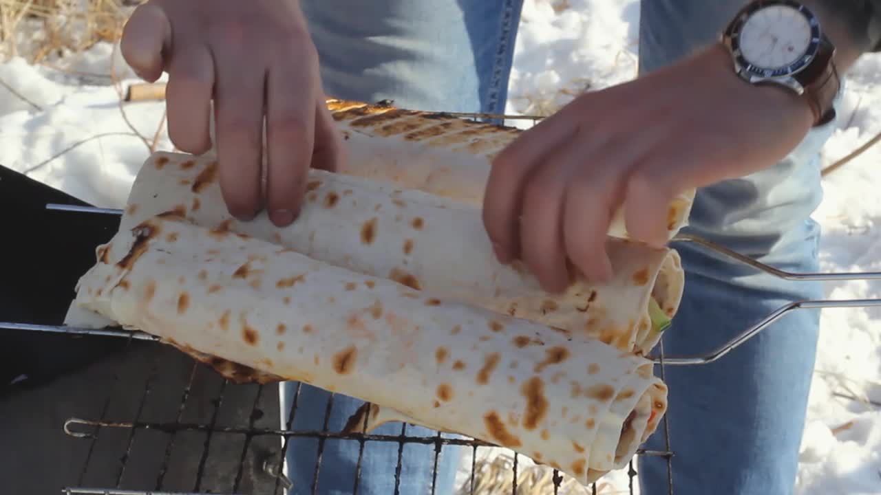 冬季野餐时，男性的手在烤架上翻转蔬菜和肉的皮塔饼卷，露营时烤制快餐视频素材