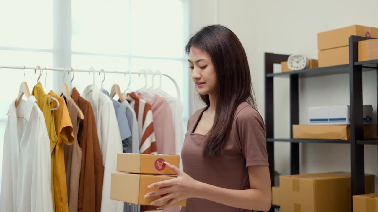 年轻快乐的亚洲妇女在时装店的老板打包衣服在包裹盒和展示给相机交付给顾客。在服装店工作的亚洲女企业家。视频素材