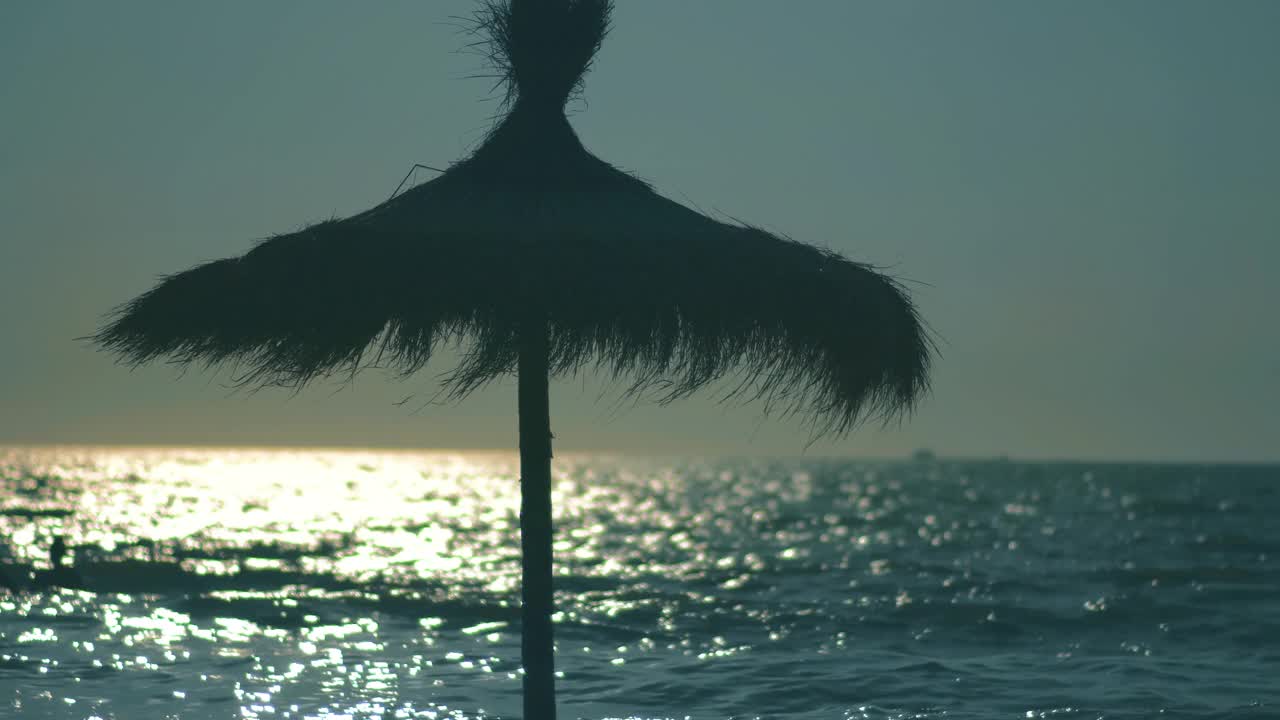 草伞在夏日的微风中挥舞着，夕阳的余晖映照在海面上。热带海滩和暑假概念视频素材