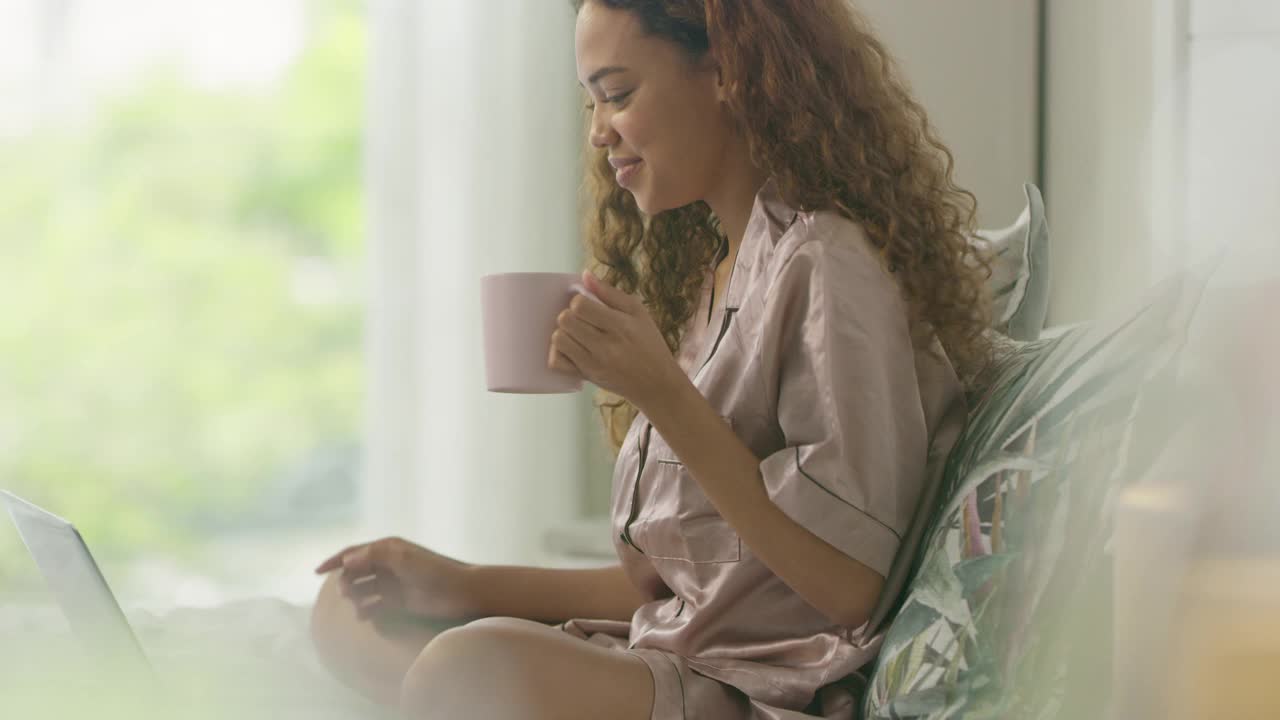 一个年轻女子在家里的床上喝咖啡时用笔记本电脑打视频电话的4k视频片段视频素材