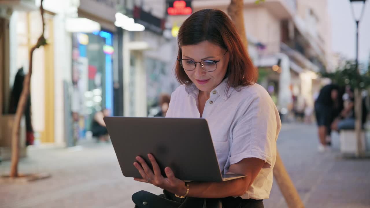 一位中年妇女坐在长椅上与一台笔记本电脑在夜晚的城市视频素材