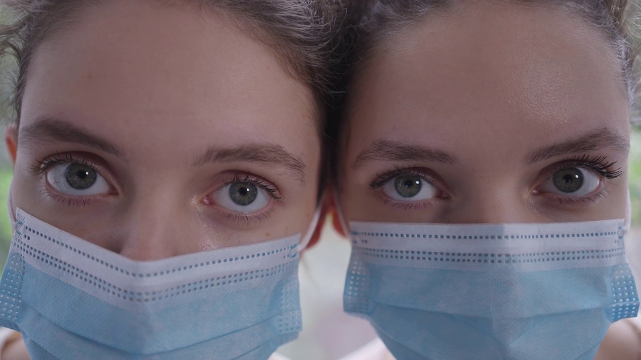 戴着冠状病毒口罩的同卵双胞胎姐妹的特写眼睛表情严肃地看着相机。苗条、年轻、美丽的白种女性因新冠疫情而被封锁的头像。视频购买