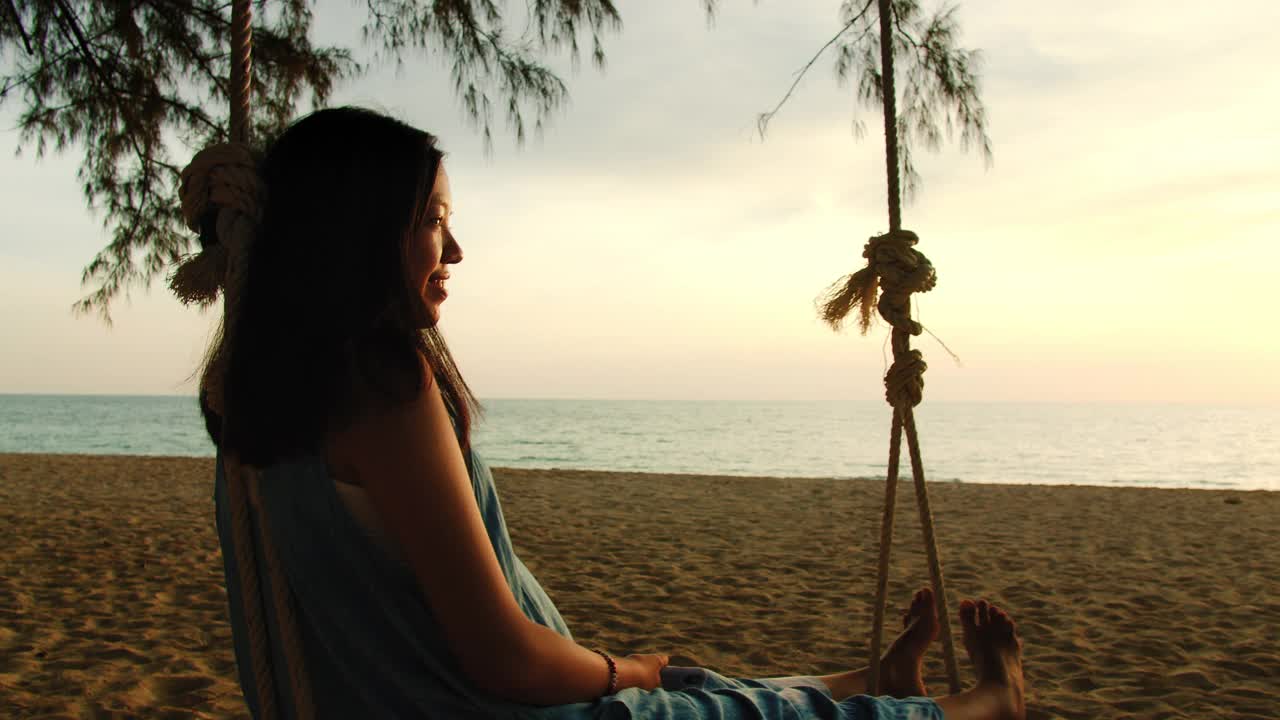 亚洲美女坐在海边的秋千上与夕阳背景在周末假期。快乐独自旅行的概念。4 k慢动作。视频素材