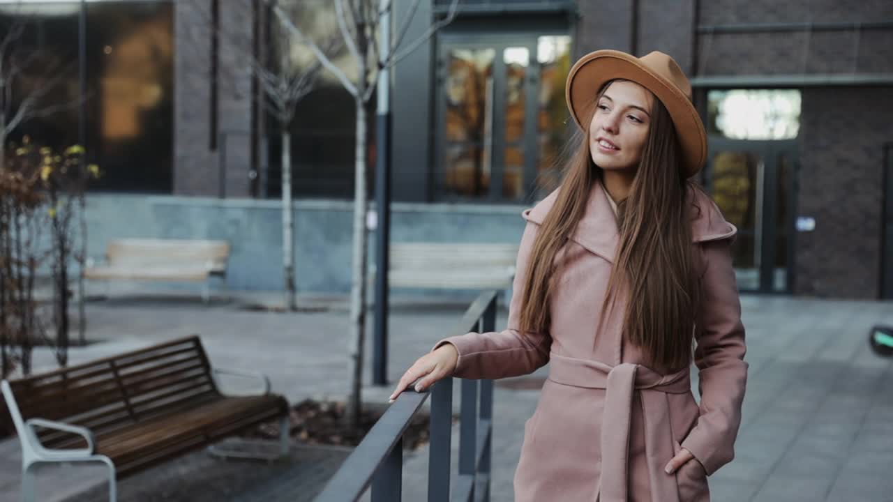 漂亮的年轻女子穿着大衣，戴着圆帽，行走在现代城市的背景下视频素材