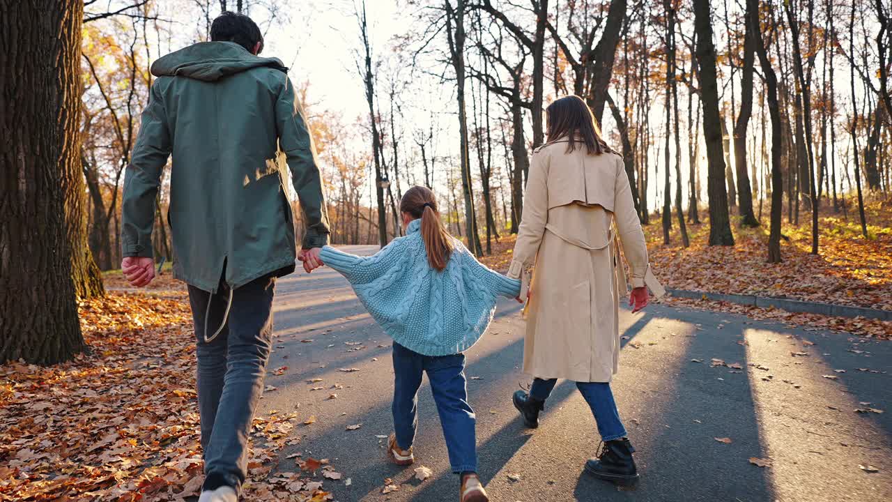 年轻的父母牵着小女儿的手，笑着玩耍，在秋天的公园里散步。特写，慢动作。跟踪多莉。后视图视频素材