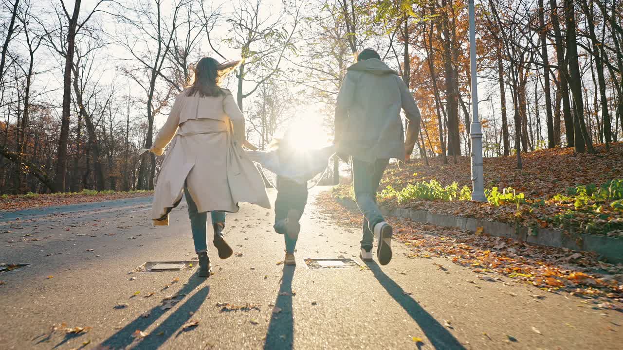 不知名的年轻父母牵着小女儿的手，在秋天公园的柏油路上玩耍奔跑。缓慢的运动。跟踪镜头，后视图视频素材