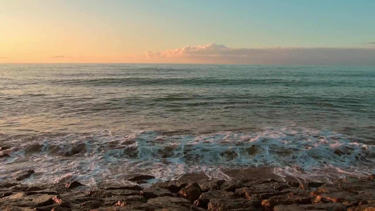 静态视图实时海浪撞击岩石在海滩上与地平线和清晰的空白蓝色橙色日落天空背景。宁静浪漫的海滩全景视频素材