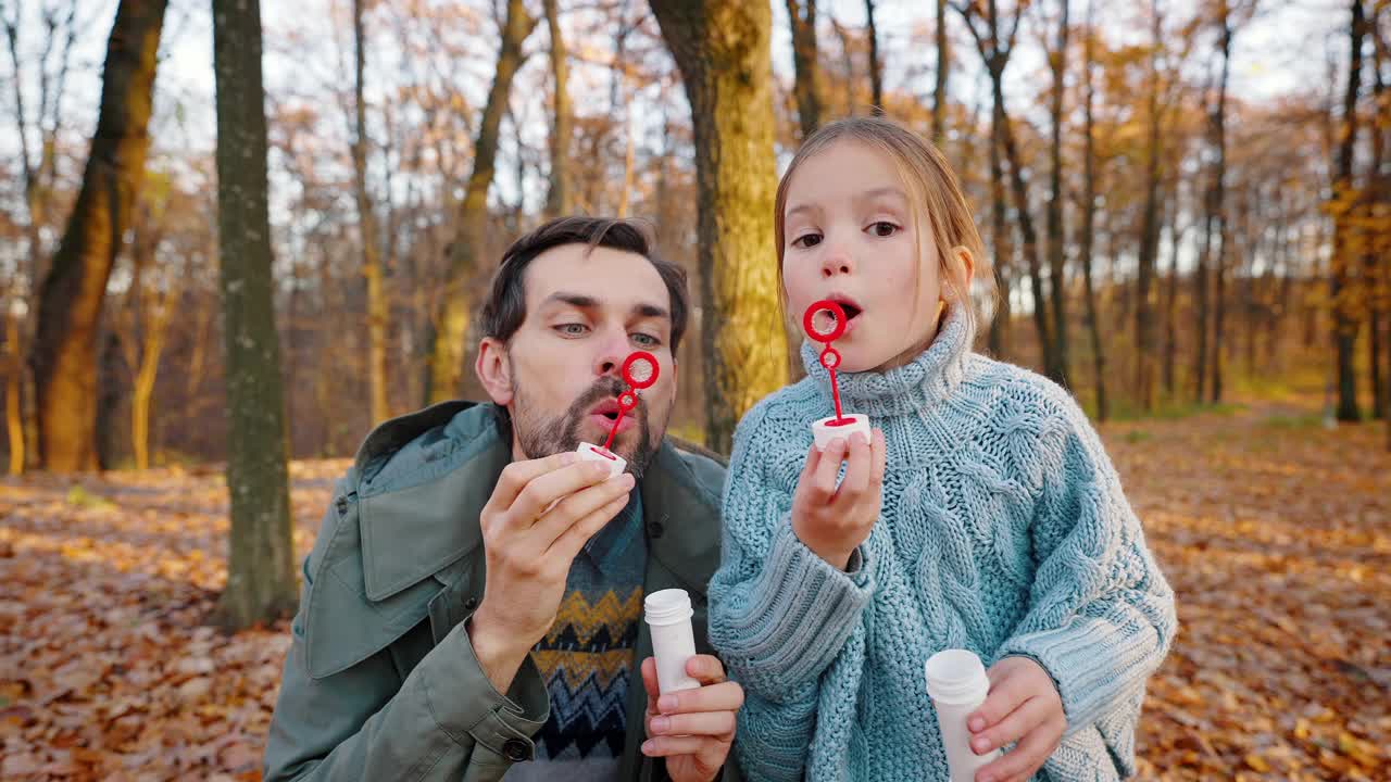 年轻的爸爸和小女儿吹着肥皂泡飞在你身上。秋天公园里的阳光明媚的一天，黄色的树叶。特写，慢动作视频素材