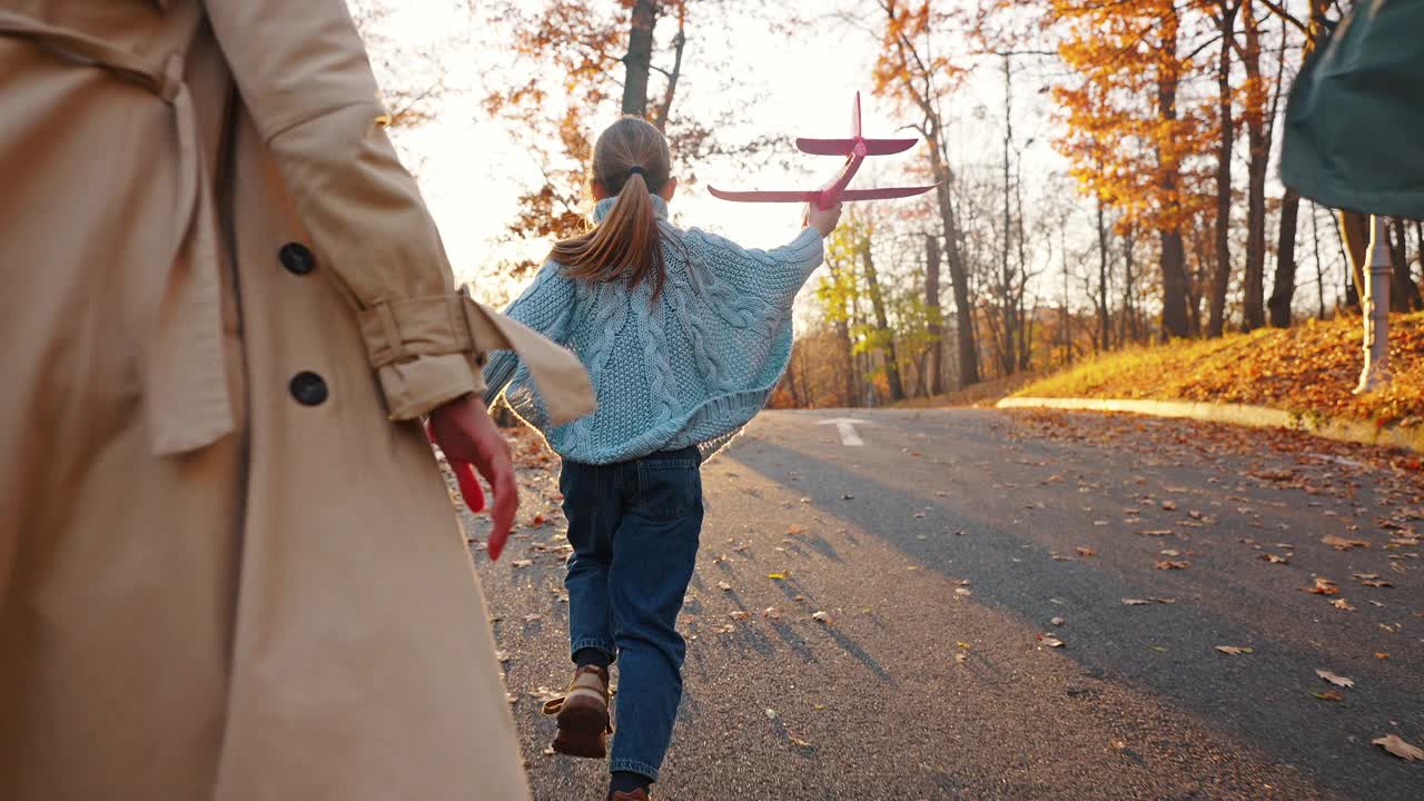 不知名的父母带着小女儿发射红色玩具飞机。秋天的公园。特写，慢动作。跟踪相机拍摄，后视图视频素材