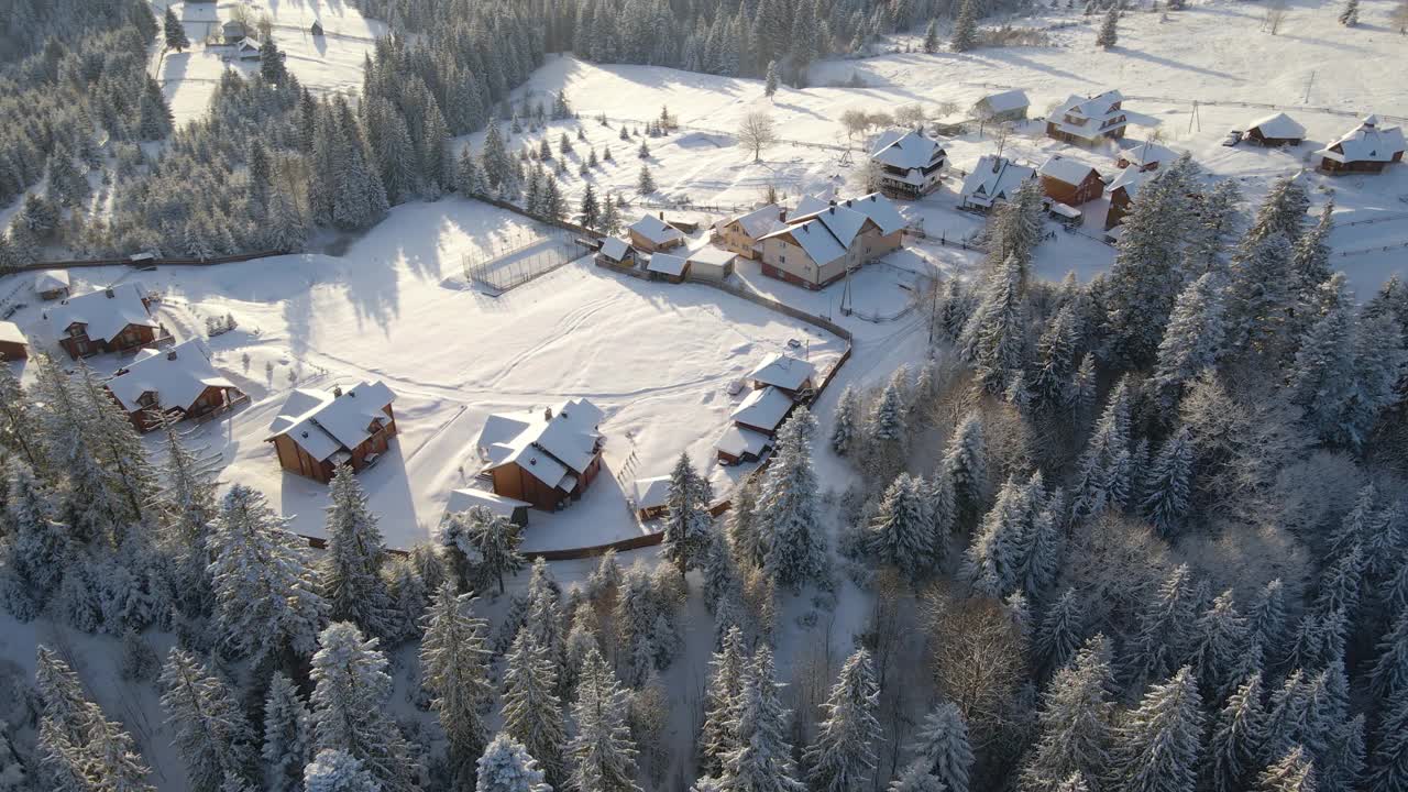 空中冬季景观与小村庄的房子之间的冰雪覆盖的森林在寒冷的山区的晚上视频素材