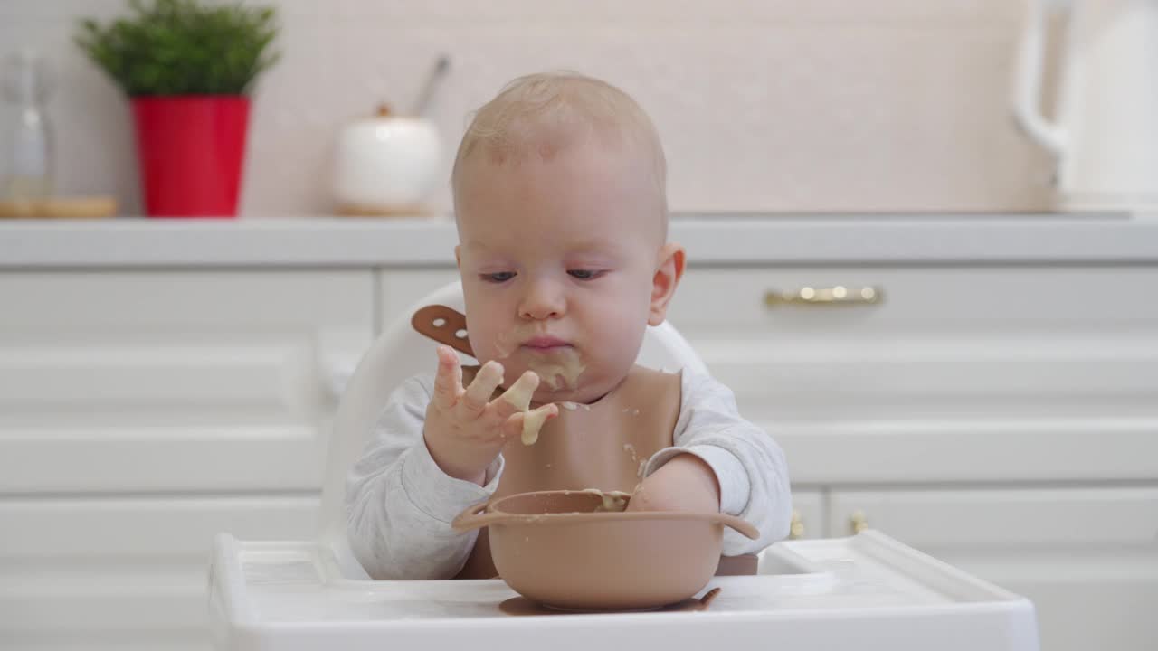 蹒跚学步的孩子学着用勺子吃饭，脸上沾着食物的孩子微笑着，婴儿坐在喂食高脚椅上，沾着粥。视频素材