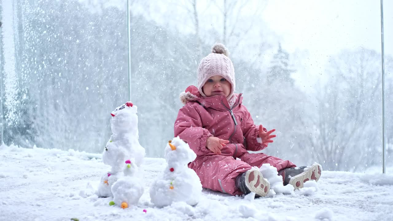 可爱的可爱的白人幼儿女婴坐在温暖的衣服在玻璃阳台阳台上玩在新鲜的白雪在美丽的冬天的一天视频素材