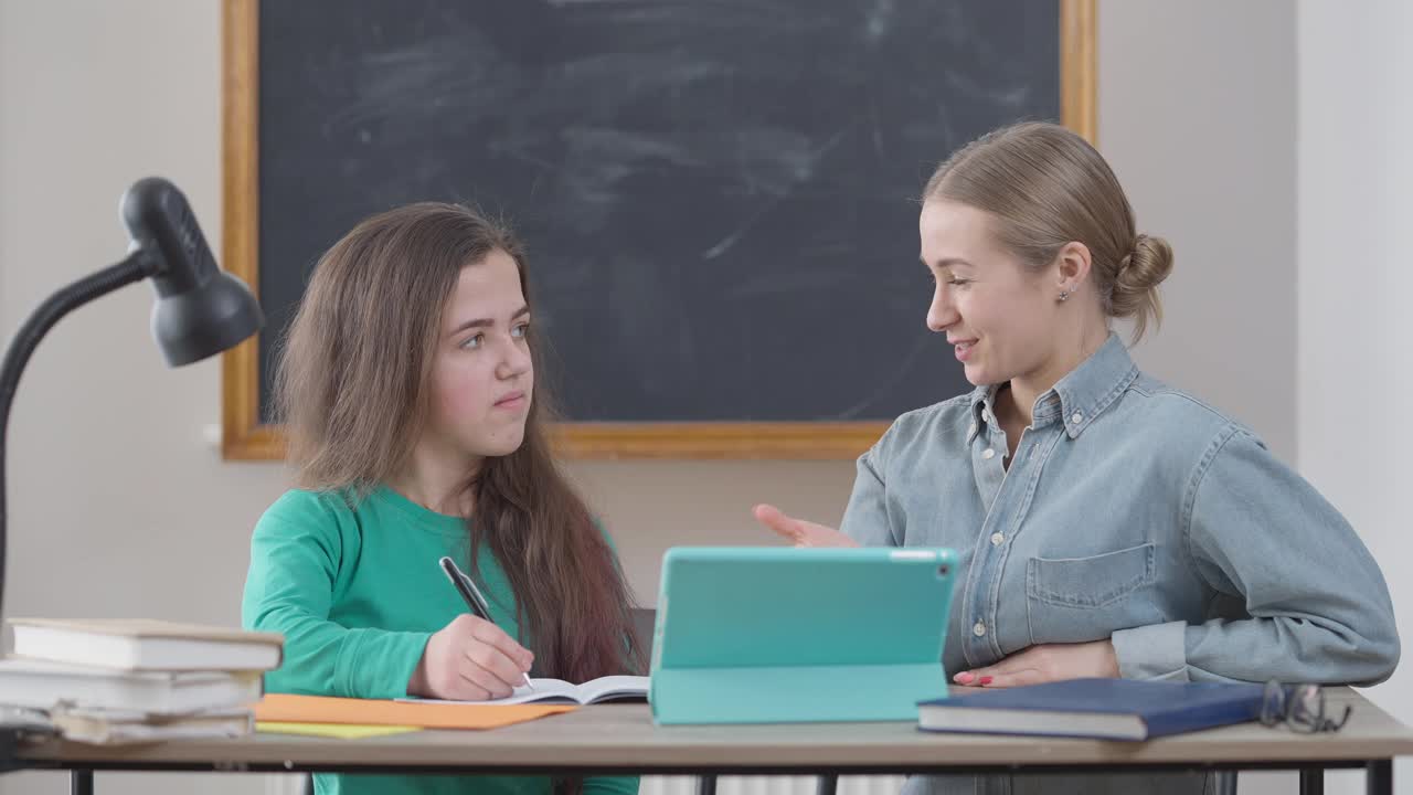 两个年轻的白人妇女坐在教室的书桌前谈论在大学学习。小人物和积极的小组成员在室内讨论研究。生活方式和智能理念。视频素材