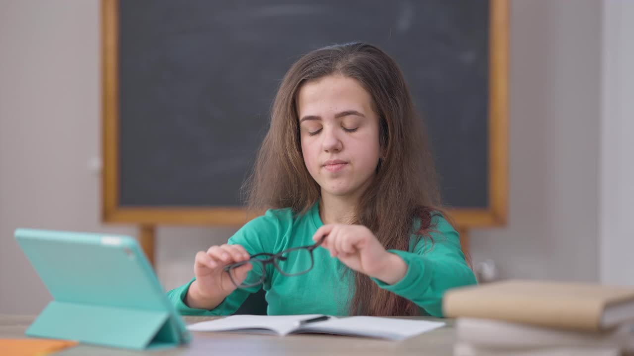 天才集中肖像白种年轻女子戴着眼镜，看着平板电脑屏幕写字，坐在书桌前。聪明自信的小个子在室内在线学习。视频素材