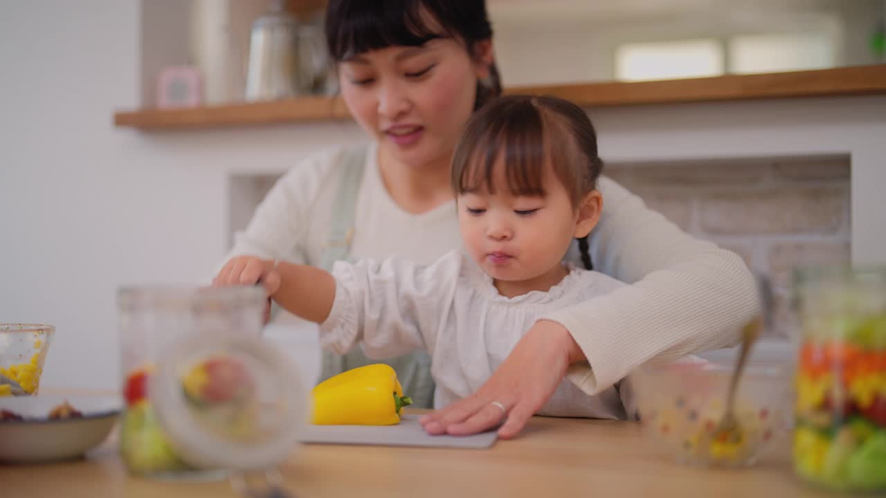 母亲和她的小女儿在家里准备沙拉。母亲帮助她的小女儿准备蔬菜，并鼓励她健康饮食视频素材