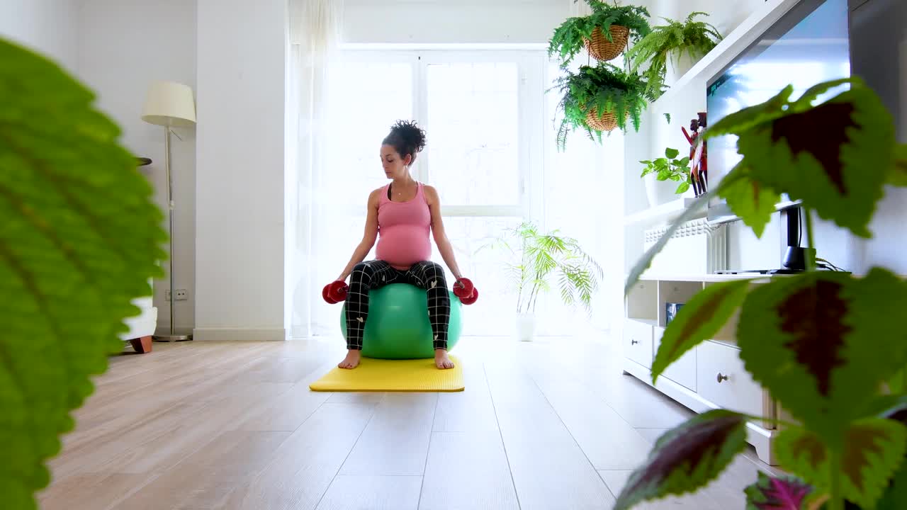 一名年轻的孕妇穿着运动服，拿着哑铃坐在家里黄色运动垫上的一个绿色健身球上。怀孕保健概念视频素材