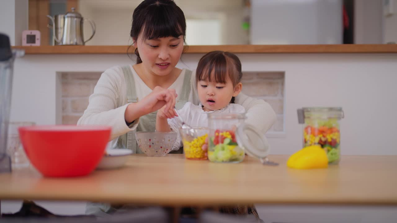 母亲和她的小女儿在家里准备沙拉。母亲帮助她的小女儿准备蔬菜，并鼓励她健康饮食视频素材