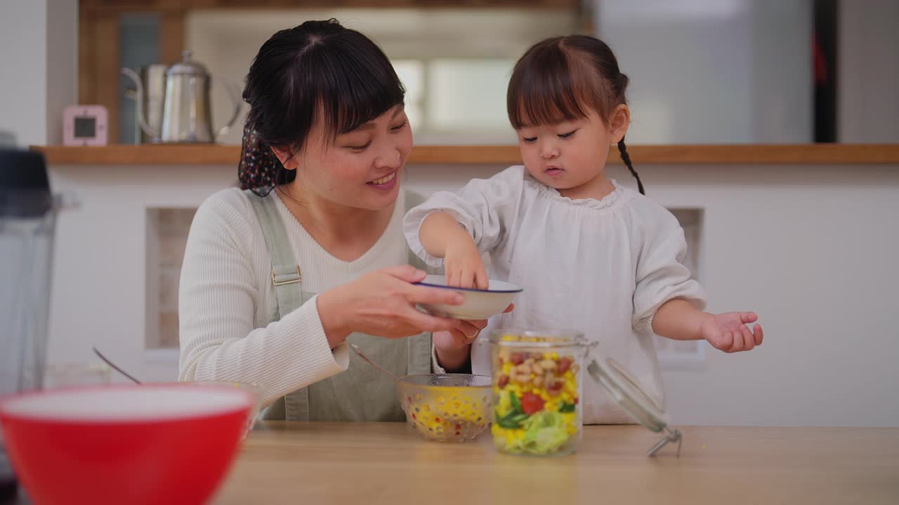 母亲和她的小女儿在家里准备沙拉。母亲帮助她的小女儿准备蔬菜，并鼓励她健康饮食视频素材