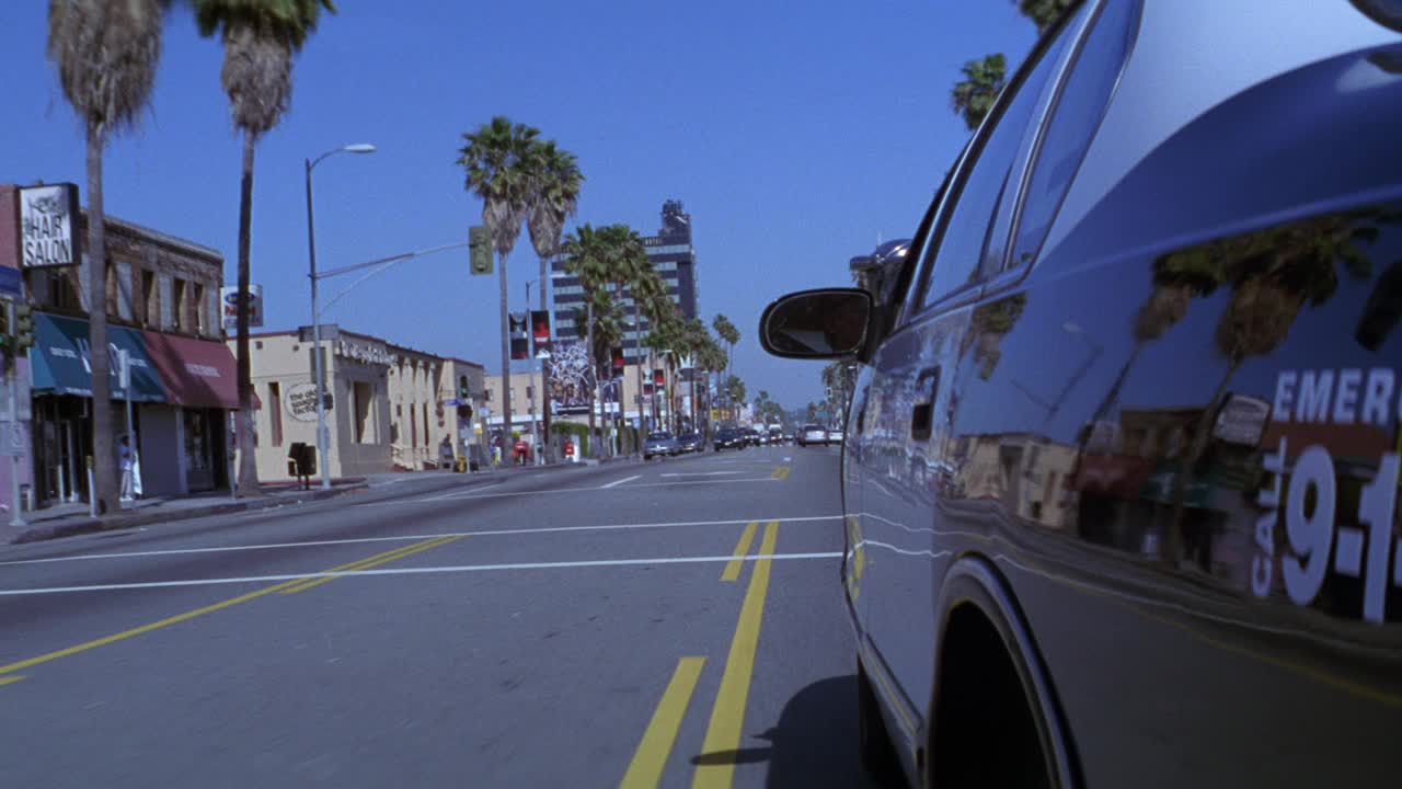 广角移动pov 3/4左前移，洛杉矶警察局警车在棕榈树林立的城市街道上超速行驶。警车急转弯，跟随向相反方向行驶的第二辆警车。多层办公楼和广告牌I视频素材