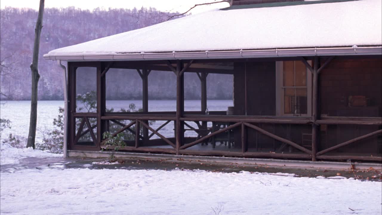 宽角小屋或带前廊的一层木湖房。背景是湖泊或河流，树木或树林。雪在地面。小雪或小雨。匹配的镜头。视频素材