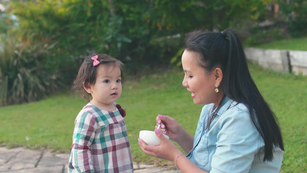 亚洲母亲在前院喂养她的混合蹒跚学步的女儿，家庭生活理念视频素材