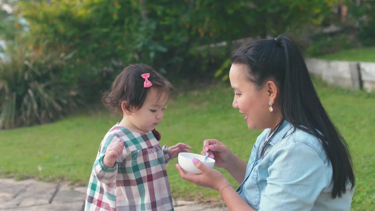 亚洲母亲在前院喂养她的混合蹒跚学步的女儿，家庭生活理念视频素材