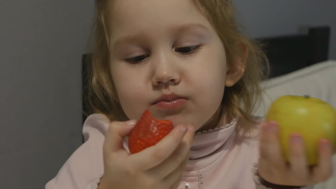 一个可爱的小女孩同时吃草莓和苹果。她依次咬了一口苹果和一个浆果。食物很好吃。健康食品的概念。4 k视频素材