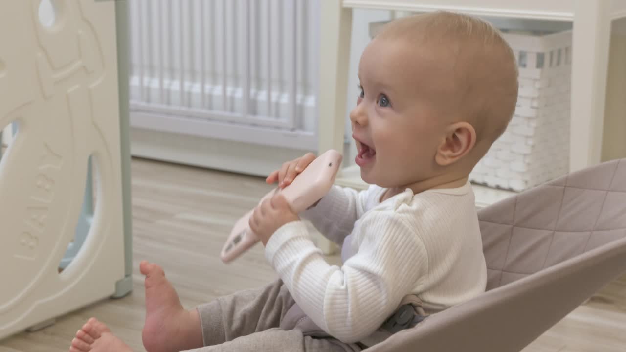 可爱的蹒跚学步的男孩坐在客厅的摇椅上，看着手机屏幕上的东西，9个月大的白人婴儿在家里使用智能手机。视频素材