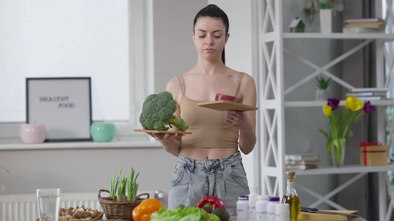 可疑的年轻女子在室内选择西兰花和肉类。深思熟虑的不确定的白人苗条美丽的千禧一代女士站在厨房决定食物。饮食和生活方式。视频素材