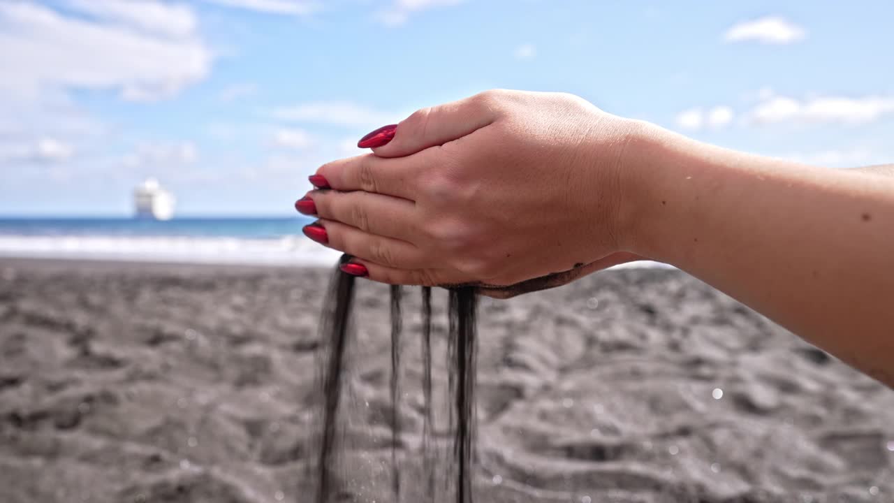 在岛屿热带海滩上，女性的手缓缓倾泻着从手指间流过的火山黑色沙子。美丽的场景，年轻女子与红色指甲玩砂砾在日落。视频素材